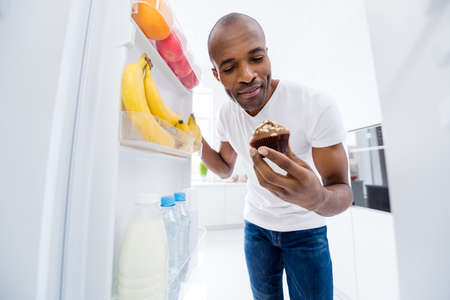 Portrait of his he nice attractive hungry guy looking in fridge taking unhealthy junk fat cupcake cake break pause regime in light white interior house kitchen indoorsの写真素材