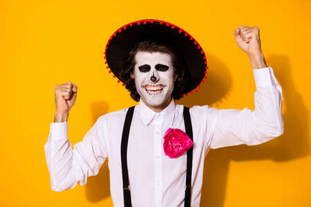 Photo of scary zombie guy raise hand fists cheerful close eyes win premium place cultural festival wear white shirt death costume sugar skull suspenders isolated yellow color backgroundの写真素材