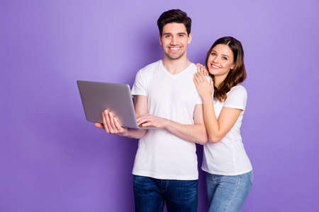 Portrait of positive couple man woman hold laptop work from home have online partnership communication wear white t-shirt casual style denim jeans isolated over violet color backgroundの写真素材