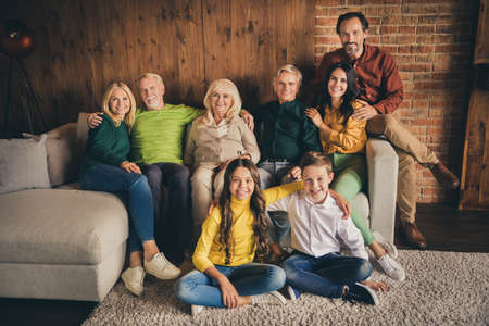 Photo of full family eight people gathering cuddle embrace satisfied good season time friendship company two little kids floor carpet sitting sofa generation in evening living room indoorsの写真素材