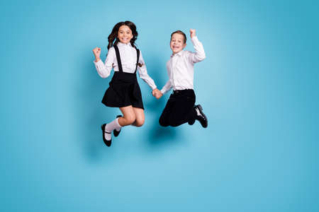 Full length photo of two opposite little girl boy schoolchildren brother sister jump hold hands have school break fun wear white shirt black pants dress isolated blue color backgroundの写真素材