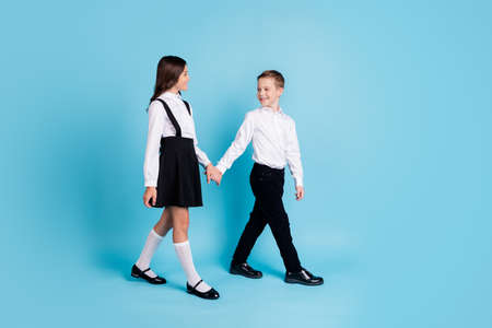 Full length profile photo of two girl boy schoolchildren smiling hold hands park walking after school wear uniform white shirt black pants dress isolated blue color backgroundの写真素材