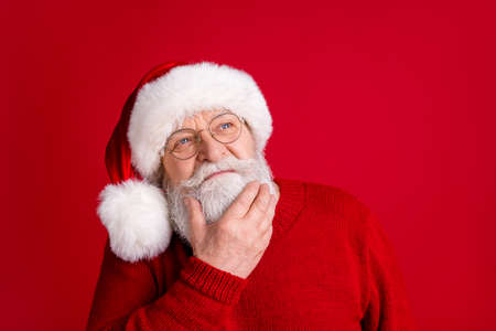 Close up portrait of unsure pensive old man in santa claus headwear look copyspace touch hand chin...