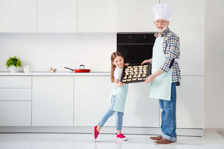 full body photo of little pretty girl granddaughter grandpa chef cap showing ready cookies on tray hobby baking together free time weekend home kitchen indoorsの写真素材