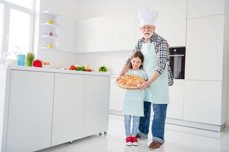 Full length body size view of cheerful grey-haired grandparent grandchild holding in hands tray baked domestic vegetarian pizza mushroom workshop help support in light white interior kitchen houseの写真素材