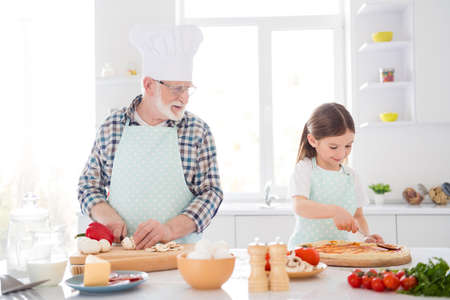 Portrait of nice cheerful cheery lovely focused grey-haired granddad grandchild cooking fresh homemade delicious meal dinner naples pizza workshop in modern light white interior kitchen houseの写真素材