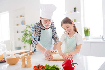 Photo of little girl granddaughter spending time with aged chef grandpa forming dough for family pizza recipe baking cooking together weekend home kitchen indoorsの写真素材
