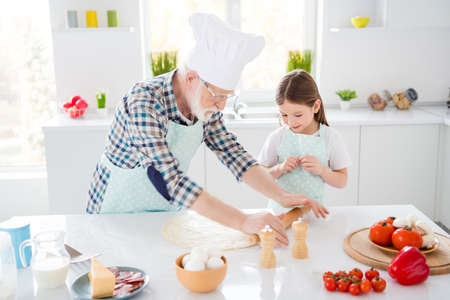 Portrait of nice cheerful grey-haired granddad teaching grandchild learning cook fresh delicious dish meal kneading dough support help assistance in modern light white interior kitchen houseの写真素材