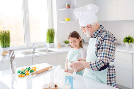 Photo of little girl granddaughter aged grandpa baking cake cookies together prepared ingredients breaking eggs make dough best friends weekend homey home kitchen indoorsの写真素材