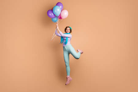 Full length photo of pretty funny lady jump high hold many air balloons raise up with wind blowing wear purple jumper green pants boots blue beret scarf isolated beige color backgroundの写真素材