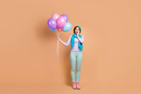 Full length photo of pretty shocked lady carry many air balloons unexpected surprise party wear lilac jumper green pants boots beret scarf isolated beige color backgroundの写真素材