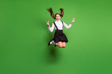 Photo portrait of crazy brunette schoolgirl screaming jumping up holding two v-signs wearing white socks isolated on vivid green colored backgroundの写真素材