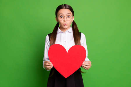 Photo portrait of schoolgirl holding big red cutout heart pouting isolated on vivid green colored...
