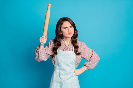 Photo of attractive crazy angry mad lady hold hands kitchen utensil rolling pin cooking cake pie want beat boyfriend for bothering wear apron dotted dress isolated blue color backgroundの写真素材