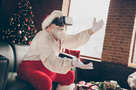 Profile side photo addicted grey beard santa claus sit couch play 3d simulation game on x-mas christmas party use modern goggles lose wear red cap headwear in house indoors evergreen treeの写真素材