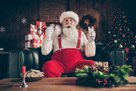 Shocked grey beard santa claus sit cozy couch watch x-mas christmas cinema film impressed drink milk glass eat gingerbread cookie wear red cap headwear in house indoors noel ornamentの写真素材