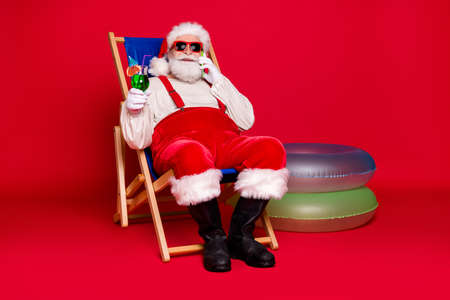 Portrait of his he handsome cheery bearded fat Santa hipster sitting in chair drinking mojito calling abroad roaming cellular rest relax chill isolated bright vivid shine vibrant red color backgroundの写真素材