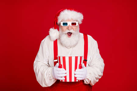 Portrait of nice handsome cheerful amazed bearded fat Santa holding in hands carton box eating popcorn watch see movie film isolated bright vivid shine vibrant red color backgroundの写真素材