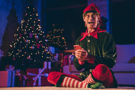 Full length photo of crazy elf sit floor hold smart phone in house indoors with christmas x-mas noel...