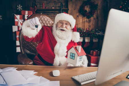 X-mas christmas lottery insurance house win concept. Grey white hair beard santa claus sit table show keys wear cap headwear lapland flat indoors noel spirit advent atmosphere ornamentの写真素材