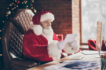 Profile side photo excited grey hair santa claus sit table impressed get wish list letter x-mas miracle fair christmas noel celebration wear red cap in house indoors with newyear decorationの写真素材