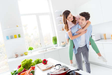 Two people funky spouses passionate man carry piggyback girl enjoy hobby weekend dinner supper lunch preparation hug in kitchen house indoorsの写真素材