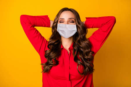 Photo of curly lady hold hands behind head eyes closed break relax meditate enjoy covid quarantine formalwear red office shirt medical mask isolated bright yellow color backgroundの写真素材