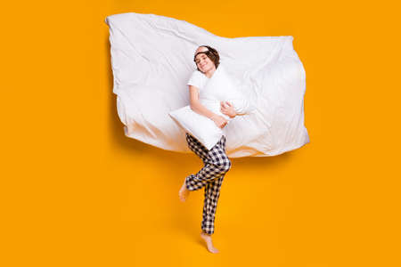 Full size photo of cheerful lady eyes closed hold hug pillow blanket flying air wake up playful energetic wear mask white t-shirt plaid pajama pants barefoot isolated yellow color backgroundの写真素材