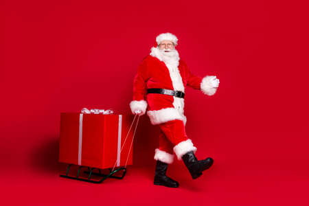 Photo portrait of man in santa claus costume pulling big wrapped gift box on sleigh isolated on vivid red colored backgroundの写真素材