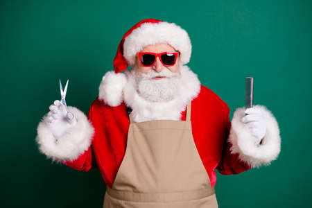 Close-up portrait of his he nice handsome bearded fat overweight Santa grandfather stylist worker wearing apron holding in hands haircut tools choosing hairdress salon isolated green color backgroundの写真素材