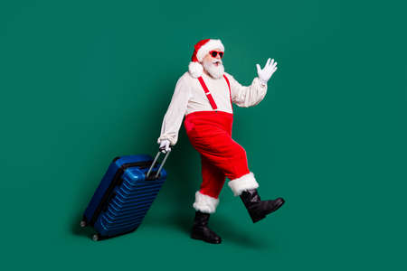 Full length body size profile side view of handsome bearded funky cheerful overweight fat Santa grandfather carrying valise air flight fly departure voyage having fun isolated green color backgroundの写真素材