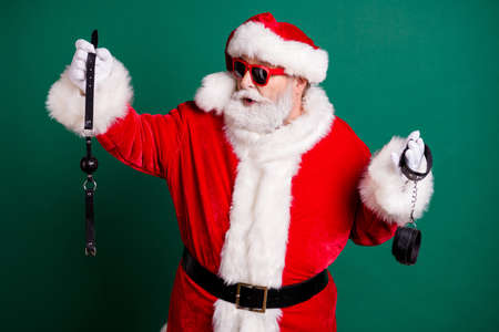 Portrait of nice attractive cheery funny Santa grandfather holding in hands choosing whip tools toys equipment passion erotica game theme party isolated over green color backgroundの写真素材