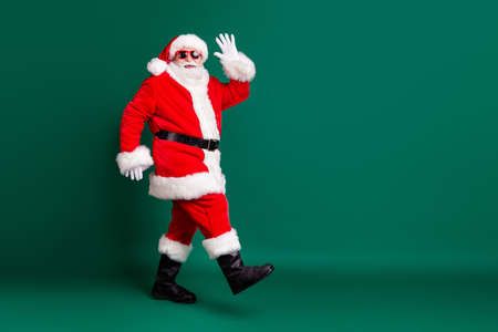 Full body size profile photo of pensioner grandpa walking mall corridors greeting shop customers wear red santa costume coat gloves sunglass headwear isolated green color backgroundの写真素材
