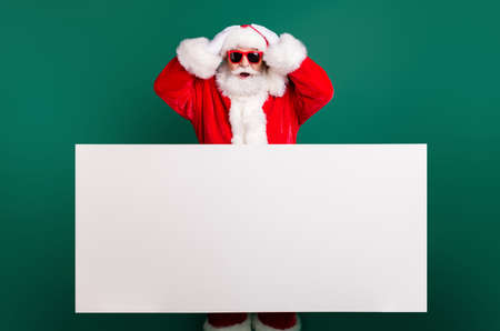Photo of retired stylish grandfather grey hair beard hands head white empty placard offer crazy discount wear red santa x-mas costume coat gloves sunglass cap isolated green color backgroundの写真素材