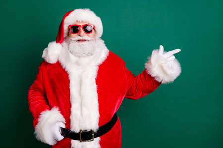 Photo of pensioner grandfather white beard smile direct finger empty space propose product wear red santa x-mas costume coat gloves sunglass belt headwear isolated green color backgroundの写真素材