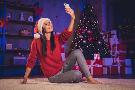 Photo portrait of woman in santa hat taking selfie sitting on floor wearing red jumper santa hat indoorsの写真素材