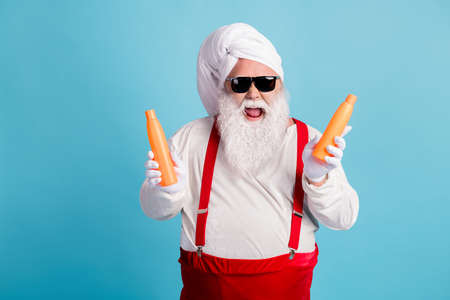 Photo of retired old fat man grey beard hold lotion bottle offer two in one price generous proposition wear santa x-mas costume towel turban suspender sunglass isolated blue color backgroundの写真素材