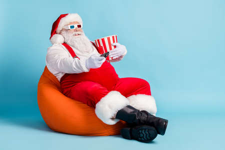 Full length photo of retired old man grey beard hold remote controll bucket watch sit orange beanbag wear red santa x-mas costume suspender 3d glasses cap boot isolated blue color backgroundの写真素材