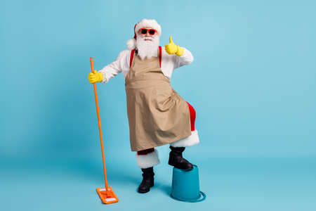 Full length photo of pensioner old man grey beard hold mop leg bucket raise thumb-up wear santa x-mas costume apron rubber glove suspender sunglass cap isolated blue color backgroundの写真素材