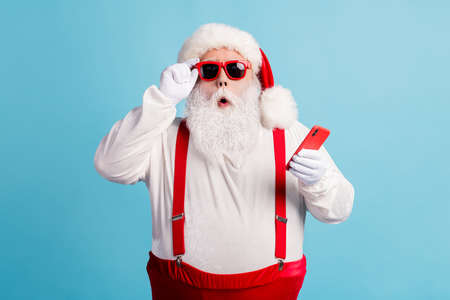 Photo of pensioner grandfather grey beard hold telephone read fake news christmas cancelled wear santa x-mas red costume suspenders sunglass white gloves cap isolated blue color backgroundの写真素材