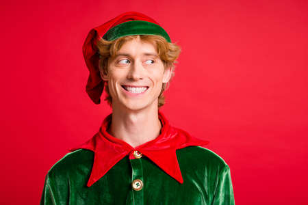 Photo of young guy look side empty space toothy smile wear santa elf green costume cap red color backgroundの写真素材