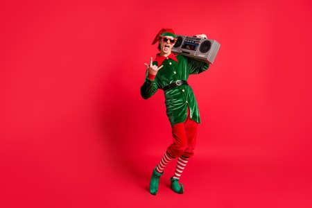 Full size photo of crazy elf hold boom box listen x-mas songs show horned symbol wear green costume isolated on red color backgroundの写真素材