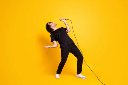 Full length profile photo of young man hold mic singer open mouth wear black t-shirt pants white sneakers spectacles isolated yellow color backgroundの写真素材