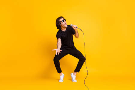 Full length photo of young man dance hold mic sing open mouth wear black t-shirt pants white sneakers sunglass isolated yellow color backgroundの写真素材