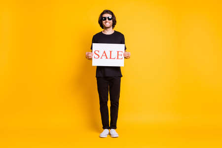 Full body size photo of young man stand hold placard sale wear black t-shirt pants white sneakers sunglass isolated yellow color backgroundの写真素材