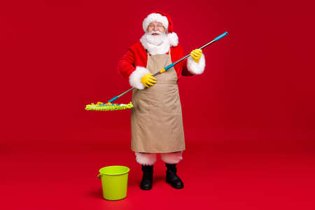 Full length photo of santa claus grey beard hold mop wear x-mas costume apron coat cap spectacles rubber gloves leather boots glasses isolated red color backgroundの写真素材