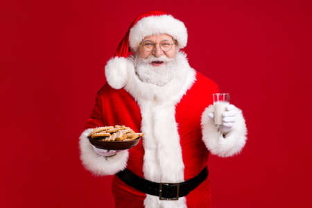 Photo of crazy funky santa claus hold x-mas tradition ginger bread cookies plate milk glass wear red costume headwear isolated over bright shine color backgroundの写真素材
