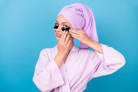 Photo of charming young lady hold use eye patches shiny smile wear violet towel turban bath robe isolated blue color backgroundの写真素材