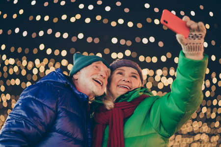 Photo of two people retired friends man woman gathering hold telephone take shoot selfie happy relationship wear coat red scarf headwear x-mas night street illumination fair outdoors outsideの写真素材