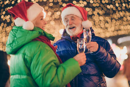 Photo of two people pensioner friends man woman meeting hold glasses drink sparkling wine laughing say magic words wear coat red scarf cap x-mas night street illumination fair outside